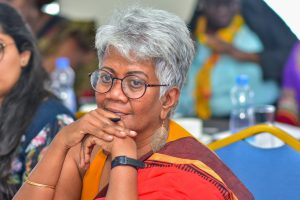 A photograph of Priyanthi at a meeting. She has short grey hair and is wearing a saree with shades of orange, red and brown. She has a thoughtful smile and is resting her chin on her hands. 