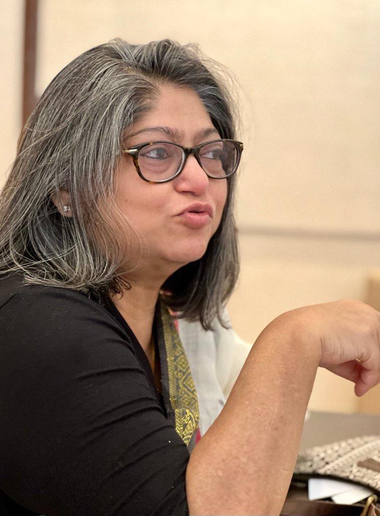 A photograph of Tulika Srivastava at a meeting. She has shoulder-length salt-and-pepper hair and is wearing a black and white saree with gold trim. Her expression is animated and she is gesturing with her right hand. 